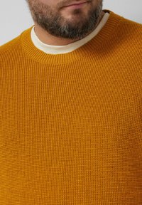 Musztardowożółty sweter dziergany o fakturowanej powierzchni i prążkowanym, okrągłym dekolcie, noszony na kremowej koszulce.