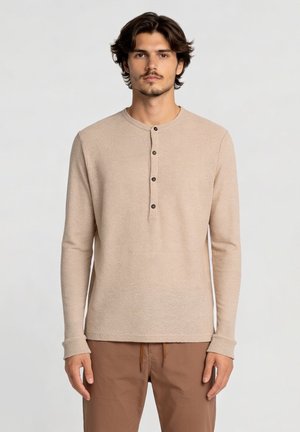 Jonge man met donker haar die een beige henley met lange mouwen en een bruine broek draagt, staand tegen een effen lichte achtergrond.