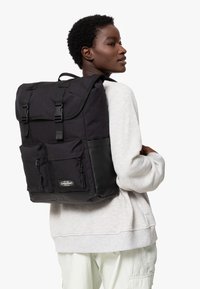 Svart ryggsäck med rulltoppdesign, dubbla framfickor med dragkedja, justerbara remmar och en glansig svart bottenpanel. Tillverkad av hållbart tyg.