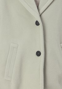 Hellgrüne Fleece-Jacke mit eingeschnittenen Revers, schwarzen Knöpfen und einer einzelnen Brusttasche. Weicher Textur, minimalistisches Design.