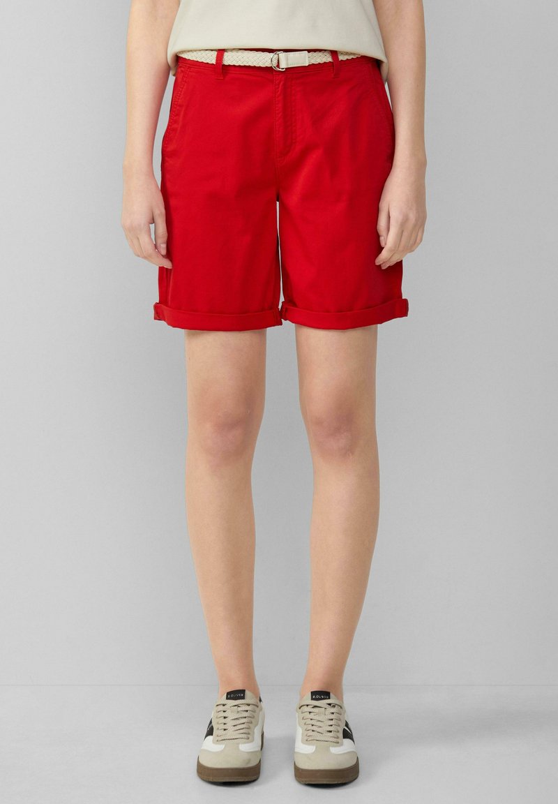 Shorts en coton rouge avec des ourlets retroussés, poches latérales et une ceinture tissée beige. Portés avec des baskets beiges et blanches.