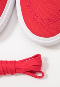 Czerwone płócienne trampki z białymi gumowymi podeszwami i szwami, z dopasowanymi czerwonymi sznurówkami leżącymi obok butów.