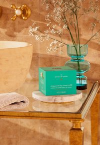 Teal låda med refill av vispad kroppskräm på en rund marmorskiva, bredvid en texturerad handduk, ett guldtablett och en klar glasvas med blommor.