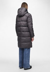 Puffjakke i mørkegrå, med hette, horisontalt quiltet design og mid-lengde silhuett. Kombinert med vide blå jeans.