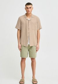 Short-sleeve, tan and brown gingham shirt with a pocket, paired with olive green shorts. Model wearing slide sandals.