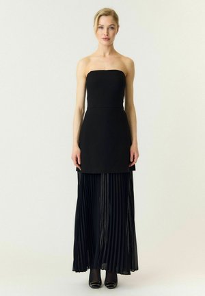 Une femme porte une robe noire bustier avec une longue jupe plissée devant un fond clair et uni.