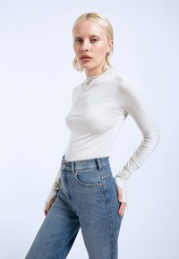 Long-sleeved, light beige top with fitted design. Paired with high-waisted, light blue denim jeans featuring side pockets. Minimalist jewelry accents.