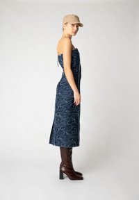 Jeanskleid in Dunkelblau mit Wellenmuster, trägerloses Design, seitliche Schlitze, kombiniert mit braunen knielangen Stiefeln und einer beigen Kappe.