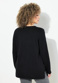 Maglione nero a maglia con maniche lunghe, scollatura rotonda e spacchi laterali. Presenta una superficie texturizzata e una vestibilità ampia, abbinato a pantaloni grigi.