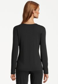 Woman with long reddish hair wearing a fitted black long-sleeve top and black pants, standing facing away against a white background.