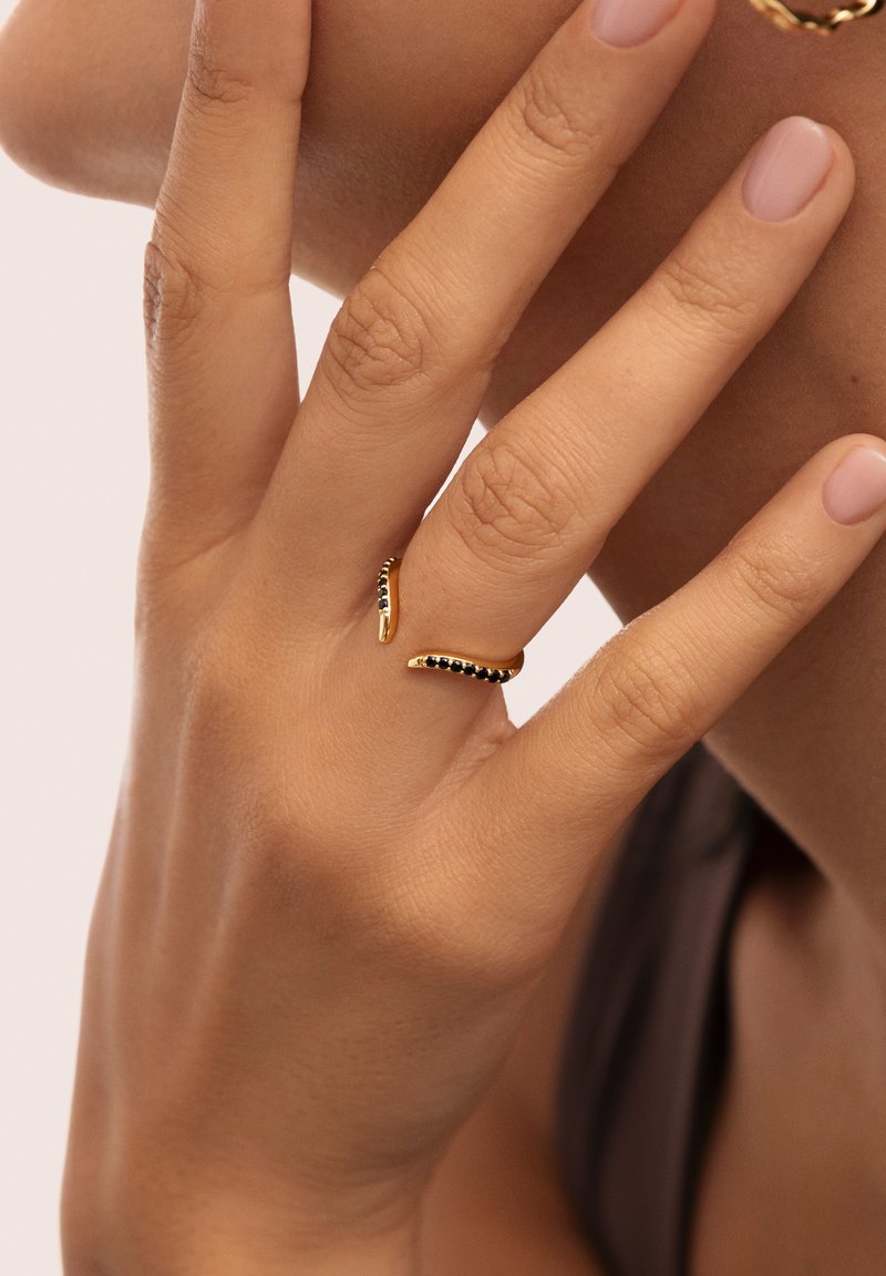 Anello d'oro con un design ondulato e accenti neri, indossato su una mano con pelle ben curata e smalto per unghie neutro.