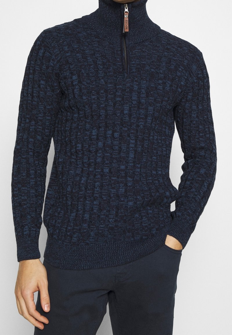 Homme portant un pull en maille texturée bleu marine avec une courte fermeture éclair et une languette en cuir, associé à un pantalon bleu marine foncé.