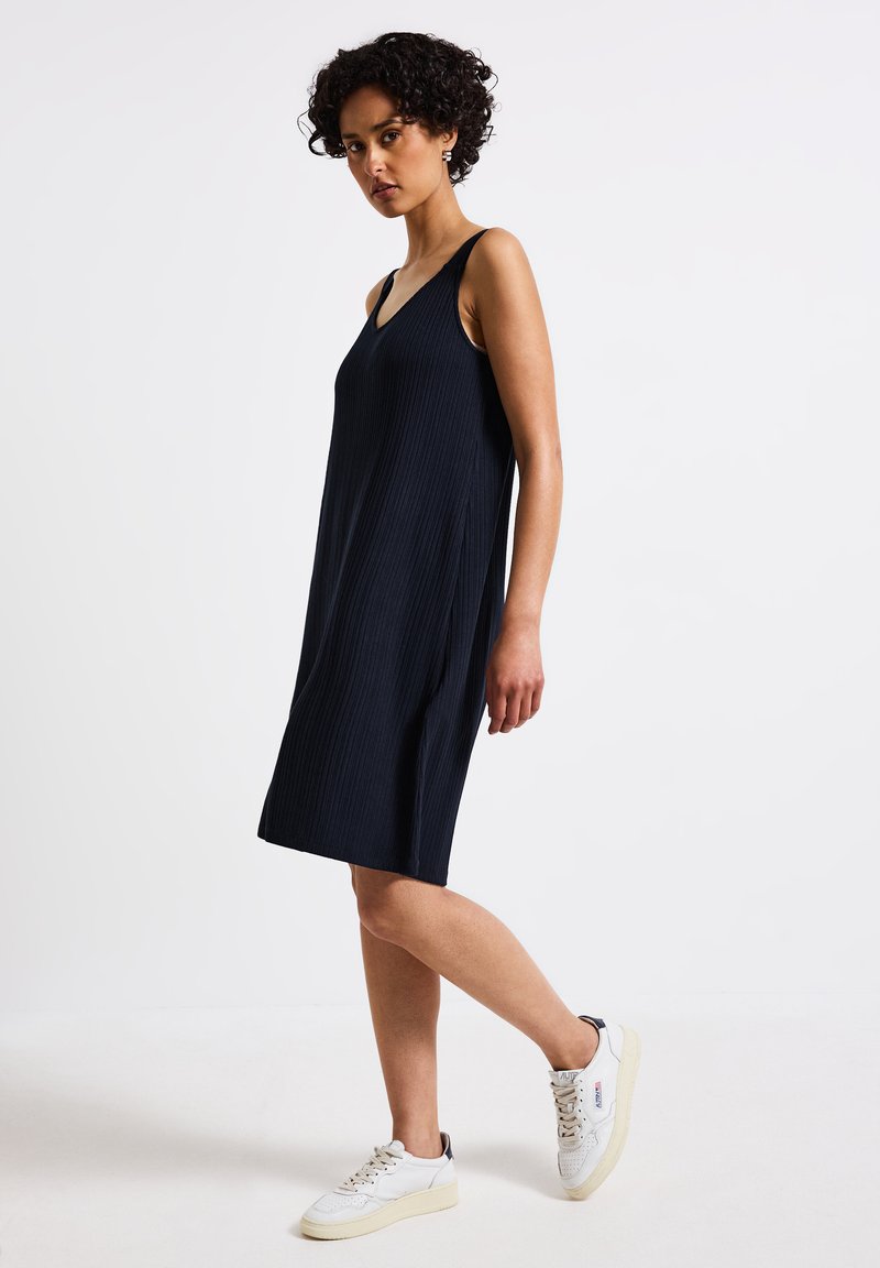 Abito nero a coste senza maniche con scollatura a V, caratterizzato da una silhouette morbida e che arriva appena sopra il ginocchio. Le sneakers bianche completano il look.