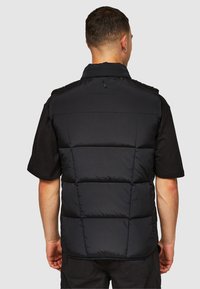 Schwarze wattierte Weste mit Kragen, versehen mit gesteppten Abschnitten und einem kleinen Logo auf der Rückseite, getragen über einem schwarzen T-Shirt.