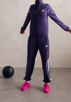 Persoon die een donkerpaarse Adidas trainingspak draagt met witte strepen en felroze sneakers, staand naast een zwarte medicijnbal.