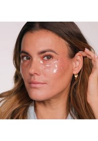 Heldere hydrogel oogpatches met roze vlekken, aangebracht onder de ogen, gedragen door een persoon met een gemiddelde huidskleur en benadrukte kenmerken.