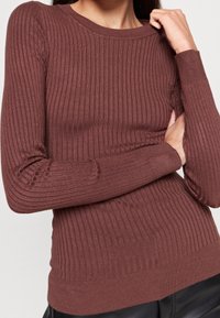 Kobieta w dopasowanym, prążkowanym, długim swetrze w kolorze brązowym z okrągłym dekoltem, z założonymi rękami i dłonią dotykającą kołnierza.