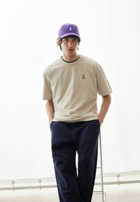 Beige katoenen T-shirt met een donker logo, gecombineerd met marineblauwe broek. Een paarse baseballpet maakt de outfit compleet, met een losse pasvorm en minimale ontwerpen.