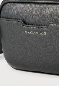Κοντινή λήψη του μπροστινού μέρους μιας μαύρης τσάντας από υφή δέρματος με το λογότυπο "Armani Exchange" σε ασημένια γράμματα.