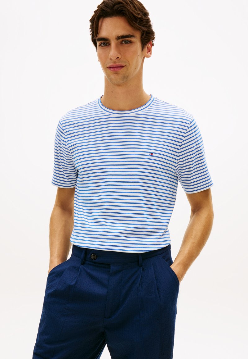 Μπλούζα με μπλε και λευκές ρίγες, κοντά μανίκια, στρογγυλή λαιμόκοψη και μικρό λογότυπο στο στήθος, συνδυασμένη με υφασμένα σκούρα μπλε παντελόνια.