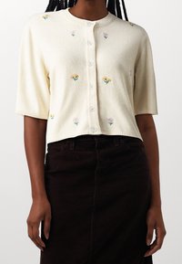 Kurzärmlige Strickjacke in Creme mit floraler Stickerei in Gelb und Lila. Verfügt über eine Knopfleiste und gerippte Abschlüsse, aus einem strukturierten Stoff gefertigt.