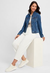 Veste en denim bleu foncé avec col classique, devant à boutons, deux poches poitrine et manches longues, associée à un pantalon blanc ajusté et des baskets crème.