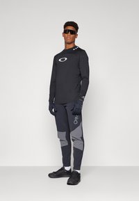 Svart langermet topp med logodetalj, kombinert med mørke bukser med grå aksenter. Svart hansker og joggesko fullfører antrekket.