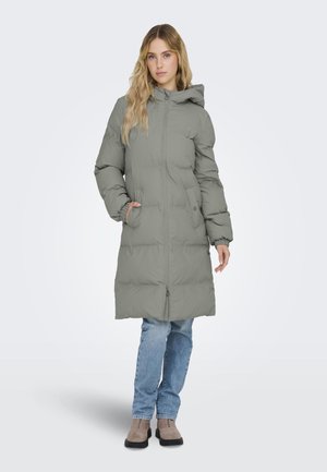 Manteau en duvet gris rembourré avec fermeture éclair à l'avant, capuche, poignets élastiques et poches latérales, associé à un jean bleu et des bottes beiges.