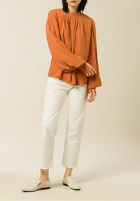 Rostorange Bluse mit langen Ärmeln, hochgeschlossen und mit gerafften Falten, kombiniert mit weißen, cropped Jeans und hellen flachen Schuhen.