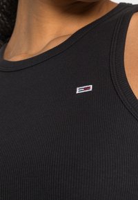 Svart ribbad linne med en liten broderad logotyp i rött, vitt och blått på bröstet. Mjuk textur med en kroppsnära silhuett.