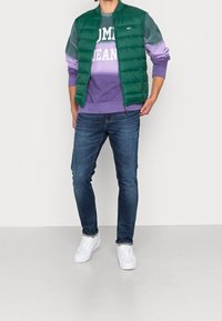 Man i grön pufferväst över lila Tommy Jeans sweatshirt, blå smala jeans och vita sneakers, som står framför en enkel bakgrund.