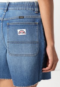 Gai zili šorti no džinsa ar saplēstu apakšmalu, aizmugurējais kabata ar Wrangler logo uzlīmi un dzeltenā diega izšuvums uz jostas.