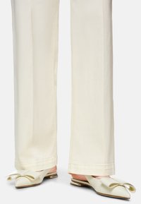 Helle, flache Mules mit einer spitzen Zehenpartie, die vorne eine große Schleifenakzentuierung und einen kleinen, glänzenden Absatz aufweisen. Kombiniert mit cremefarbenen Hosen.