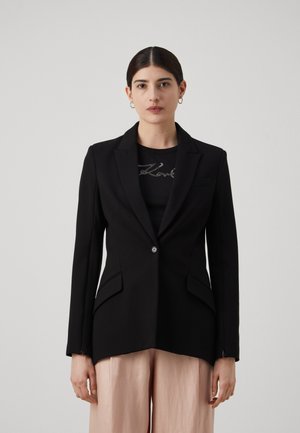 Black blazer with notched lapels, single button closure, and two front pockets, paired with a black top and light pink wide-leg trousers.
