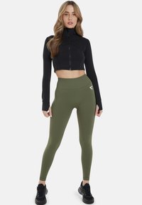 Mustat lyhyet zip-up-takit pitkillä hihoilla, yhdistettynä oliivinvihreisiin korkeavyötäröisiin legginsseihin, joissa on huomaamaton logotiedosto.