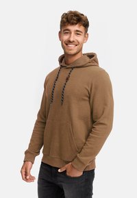 Brun sweatshirt med hætte og ribbet tekstur, kontrastfarvede marineblå snore og en frontlomme. Båret med sorte bukser.