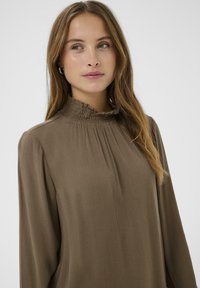 Ruskea pitkähiha blouse, jossa on poimutuksia ja smockattu korkea kaulus, valmistettu teksturoidusta kankaasta. Kauluksessa on ribbiteksturointia.