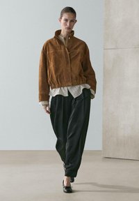 Vrouw die loopt, gekleed in een bruine suède jas, gestreept shirt, losse zwarte broek en zwarte ballerina's, voor een effen achtergrond binnenshuis.