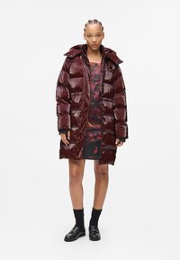 Burgundy padded coat with a glossy finish, featuring a high collar, zip closure, and large quilted sections. Worn over a printed dress.