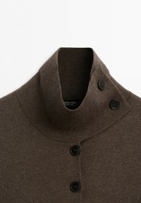 Brun stickad tröja med hög krage och tre svarta knappar vid halsen och detaljer för frontstängning.