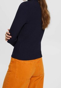 Marineblauer gerippter Rollkragenpullover kombiniert mit orangefarbenen Hosen mit weitem Bein, ausgestattet mit zwei Gesäßtaschen und einem strukturierten Stoff für zusätzliche Optik.