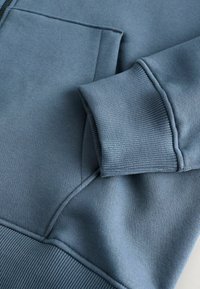 Gros plan sur un tissu de sweat-shirt bleu avec un poignet côtelé et une poche, présentant des détails de couture visibles.