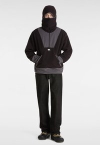 Schwarzer und grauer Fleece-Pullover mit hohem Kragen und Reißverschluss, ausgestattet mit großen Fronttaschen. Das Modell trägt dunkle Hosen und Schuhe mit Akzenten.