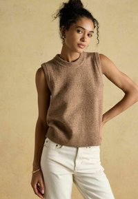 Brauner ärmelloser Strickpullover mit einem gerippten Hals- und Saumabschluss, kombiniert mit weißen Jeans. Der Stoff wirkt weich mit einer strukturierten Oberfläche.