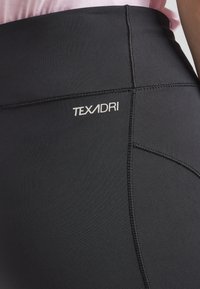 Leggings neri con una superficie liscia, caratterizzati da un logo bianco "TEXADRI" e dettagli di cucitura a contrasto. La vista ravvicinata si concentra sulla vita.