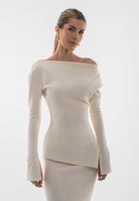 Top a spalle scoperte con maniche lunghe in tessuto a coste color crema, caratterizzato da una silhouette aderente e un orlo leggermente svasato. Texture liscia.