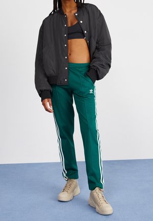 Personne portant un pantalon de survêtement Adidas vert foncé avec des rayures blanches, des baskets beige, un soutien-gorge de sport noir et une veste bomber noire sur un fond uni.