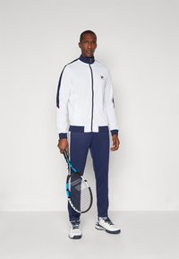 Men's athletic tracksuit in white and navy, featuring a zip-up jacket with color-blocked sleeves and matching pants, holding a tennis racket.