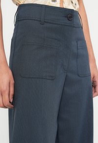 Pantalon bleu foncé texturé à taille haute, avec deux poches avant et une fermeture à un bouton. Motif à fines rayures subtil visibles.