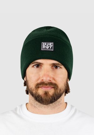 Green knitted beanie with a ribbed texture and a square black label featuring white text. Worn on a person against a neutral background.